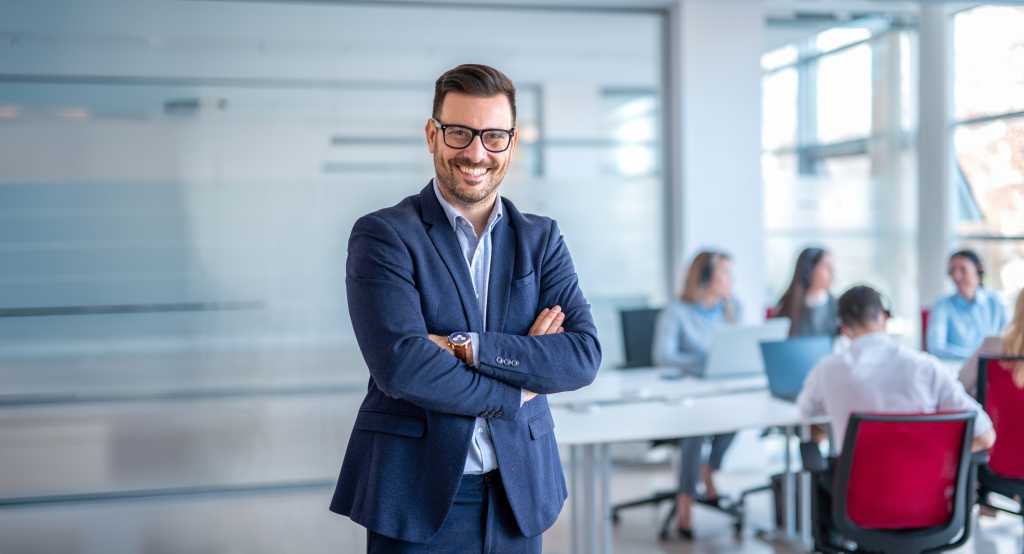 Successful businessman in formal wear standing with folded arms in modern coworking office and looking looking at camera.