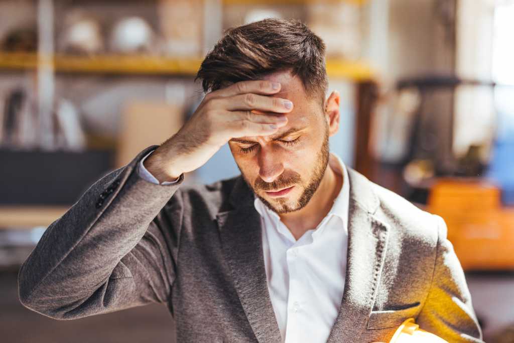 A businessman, visibly stressed, holds his head in a busy industrial environment, reflecting work tension and pressure.