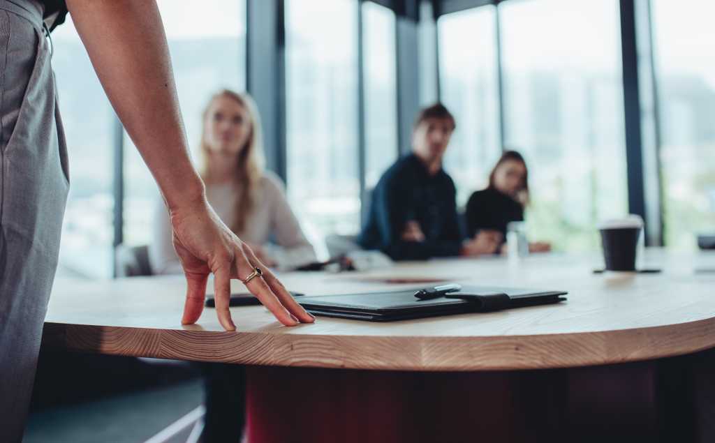 Female manager standing at table and addressing her team sitting in meeting room. Focus on hand of businesswoman in table while talking with colleagues in boardroom.